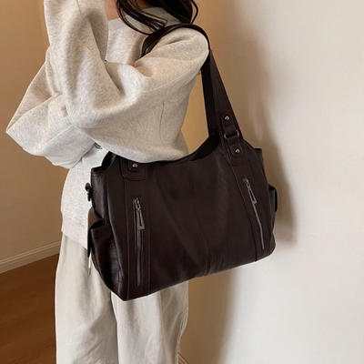 Sac à main en cuir marron porté par une femme en pull gris et pantalon beige, design moderne avec fermetures éclair, mode élégante et tendance.