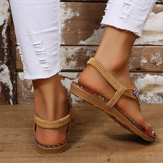 Sandales bohèmes multicolores sur pieds féminins, portées avec un jean blanc déchiré, posées sur un sol en bois rustique. Mode estivale décontractée.