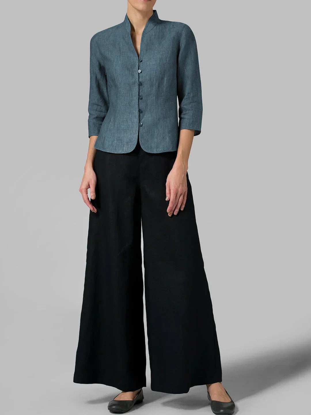 Femme portant une veste en lin bleu à manches trois-quarts et un pantalon large noir, posant sur fond gris. Mode élégante et minimaliste.