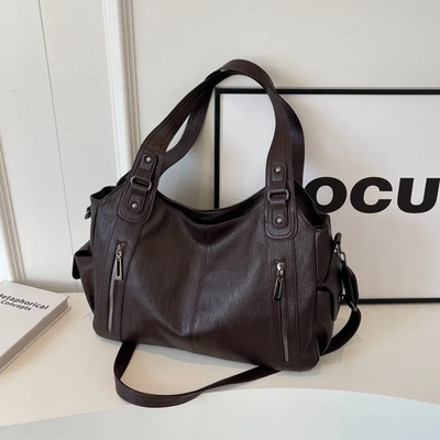 Sac à main en cuir marron foncé avec bandoulière, poches zippées, posé sur une table blanche. Accessoire mode élégant et pratique pour femme.
