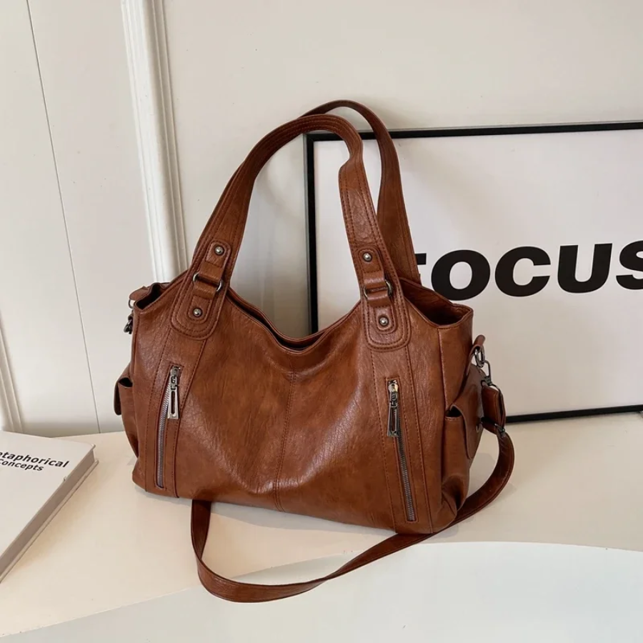 Sac à main en cuir marron avec bandoulières, poches zippées, posé sur une table blanche près d'un cadre et d'un livre. Mode, accessoires, élégance.