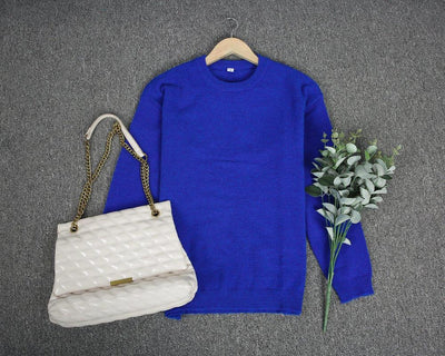 Pull bleu en laine sur cintre, sac à main beige avec chaîne dorée, bouquet de feuilles vertes, posés sur tapis gris. Mode automne-hiver élégante.