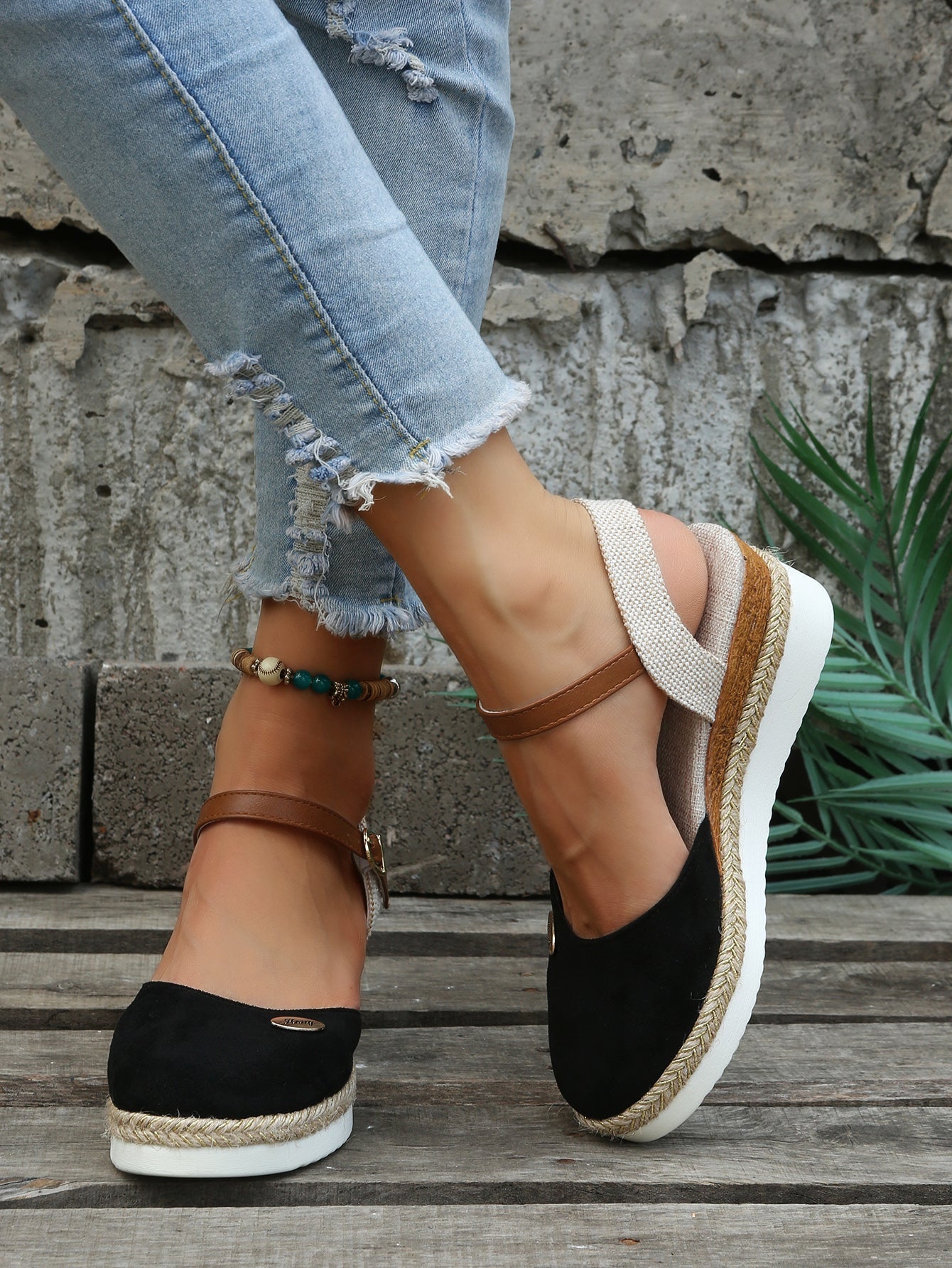 Chaussures compensées noires en daim avec bride marron, portées avec un jean déchiré, posées sur un sol en bois devant un mur en béton.