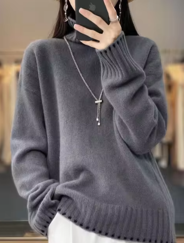Femme portant un pull gris en laine avec un collier pendentif, tenant un smartphone. Mode automne-hiver, style décontracté et élégant.