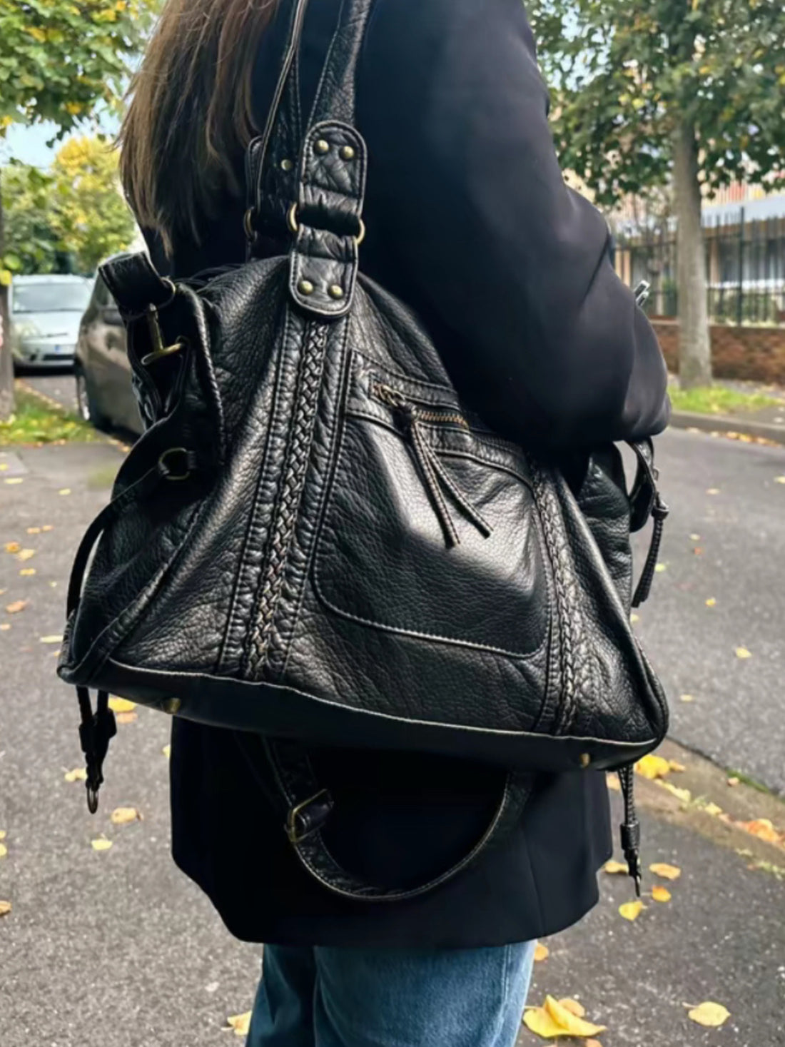 Sac à main en cuir noir porté par une femme, avec détails de couture et poches zippées, sur fond de rue automnale avec feuilles jaunes.
