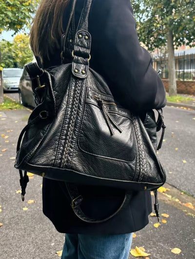Sac à main en cuir noir porté par une femme, avec détails de couture et poches zippées, sur fond de rue automnale avec feuilles jaunes.