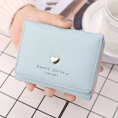 Portefeuille bleu pastel en cuir synthétique avec un motif de cœur doré, tenu par une main sur une table à carreaux. Accessoire mode élégant.