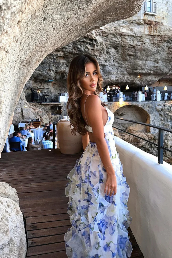 Femme en robe longue à fleurs bleues et blanches, posant dans un restaurant en falaise avec vue sur la mer, ambiance élégante et estivale.