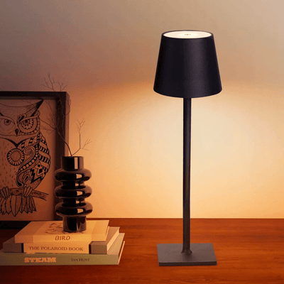 Lampe de table moderne avec abat-jour noir, posée sur une table en bois, à côté de livres empilés et d'un vase noir, éclairant un décor intérieur.
