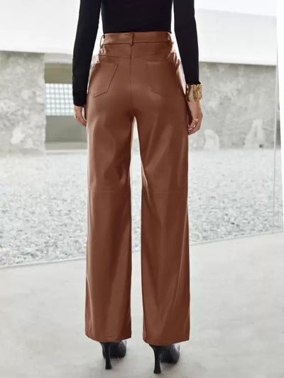 Femme portant un pantalon en cuir marron, vue de dos, avec des talons noirs et un haut noir, dans un environnement moderne et minimaliste.