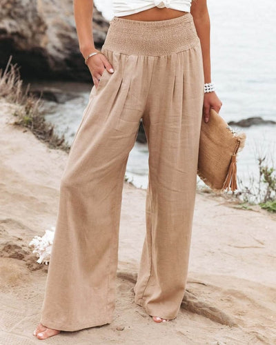 Pantalon large beige en lin pour femme, taille haute, sur une plage. Mode estivale, tenue décontractée, accessoire sac en paille.