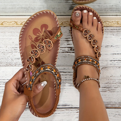 Sandales bohèmes marron avec motifs floraux et perles colorées, portées sur pieds féminins avec pédicure blanche, sur fond de bois blanc.