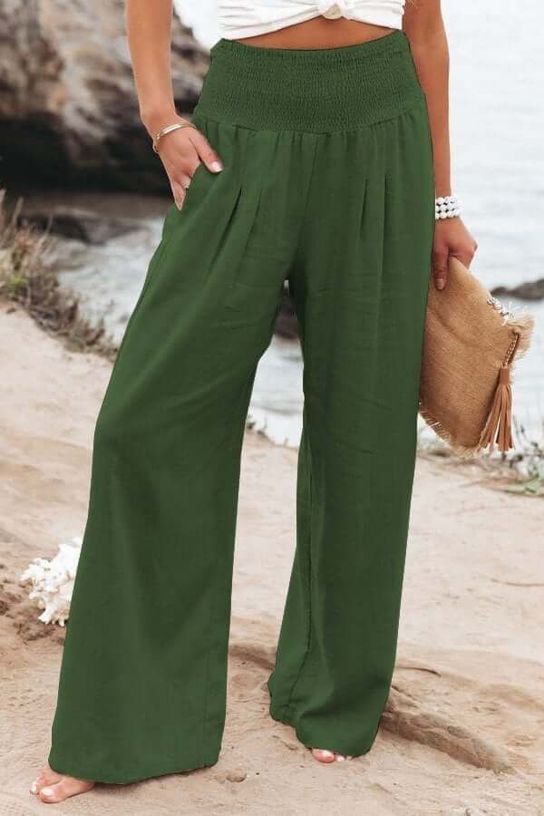 Femme en pantalon large vert, bord de mer, tenant un sac en paille. Mode estivale, tenue décontractée, plage, style bohème chic.