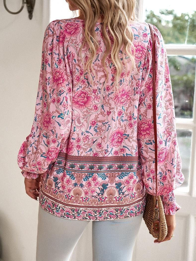 Femme portant une blouse à motifs floraux roses, manches longues, avec un sac en osier, debout devant une fenêtre. Mode féminine, style bohème.