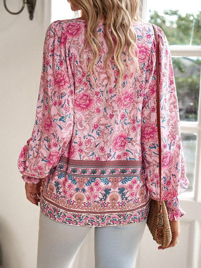 Femme portant une blouse à motifs floraux roses, manches longues, avec un sac en osier, debout devant une fenêtre. Mode féminine, style bohème.