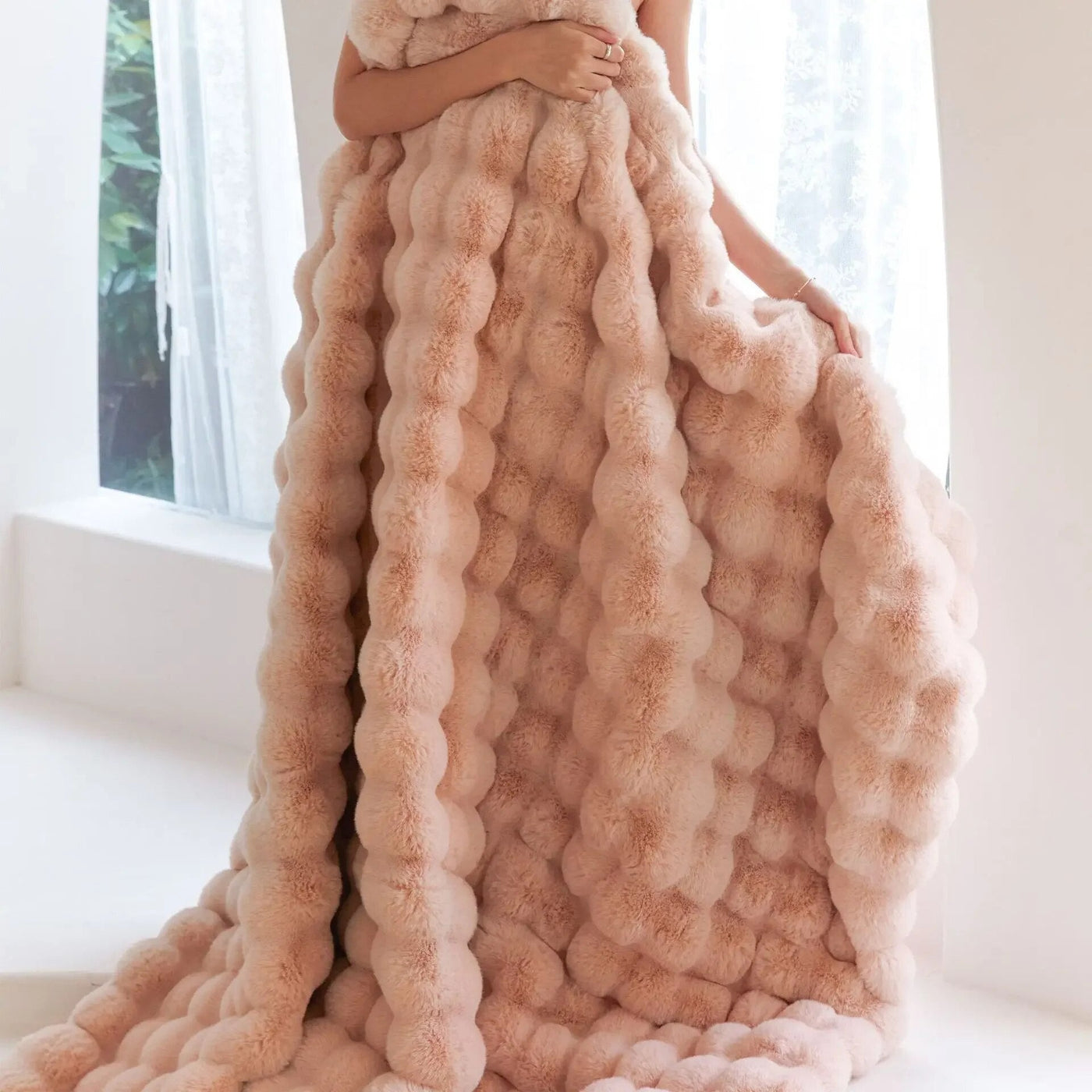 Femme enveloppée dans une couverture en fausse fourrure rose, texture moelleuse, intérieur lumineux, rideaux blancs, ambiance cosy et élégante.
