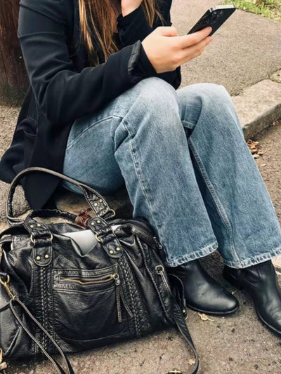 Femme assise sur un trottoir, portant un jean et des bottes noires, utilisant un smartphone. Sac à main en cuir noir à ses côtés.
