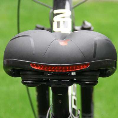 Vue arrière d'une selle de vélo ergonomique noire avec lumière LED rouge intégrée, posée sur un vélo noir, sur fond de pelouse verte.