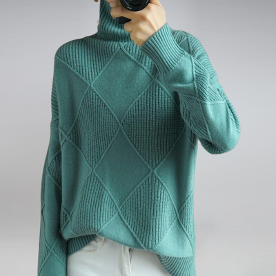 Pull en maille turquoise à col roulé, motif losanges, manches longues, porté par une personne tenant un appareil photo. Mode automne-hiver tendance.