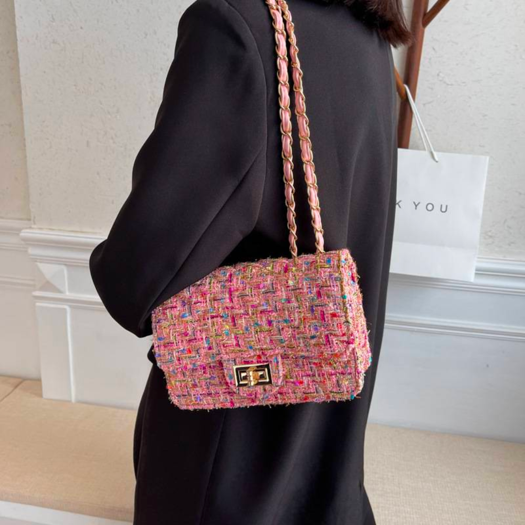 Sac à main en tweed multicolore porté par une femme en manteau noir, avec chaîne dorée, devant un mur blanc. Accessoire mode élégant et tendance.