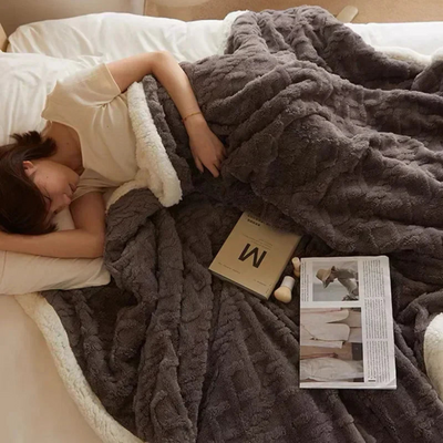 Femme dormant sous une couverture en fausse fourrure grise sur un lit, avec un livre et un magazine ouverts à côté. Confort, détente, literie douillette.