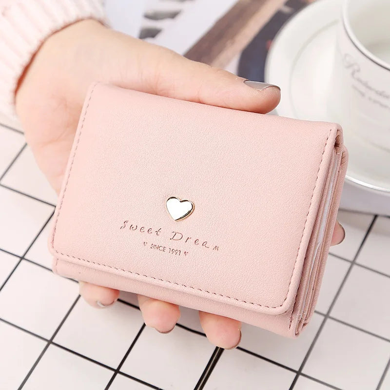 Portefeuille rose en cuir avec un motif de cœur doré, tenu par une main. Accessoire élégant pour femmes, idéal pour la mode et le rangement.