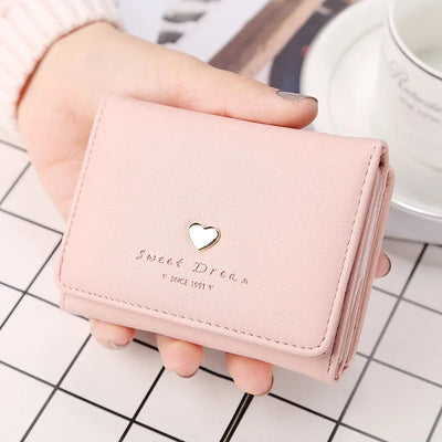 Portefeuille rose en cuir avec un motif de cœur doré, tenu par une main. Accessoire élégant pour femmes, idéal pour la mode et le rangement.