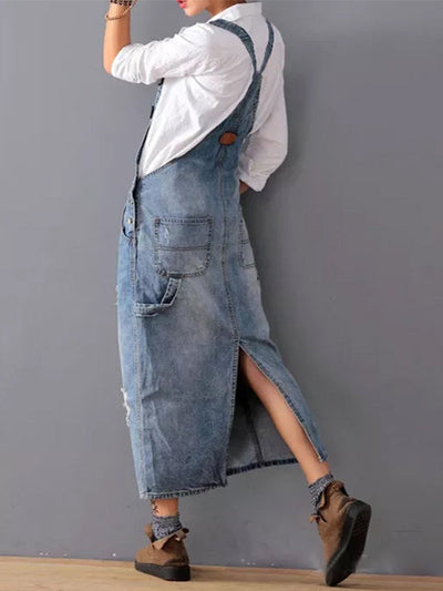 Femme portant une robe salopette en jean bleu avec chemise blanche, chaussures marron, posant contre un mur gris. Mode décontractée, style urbain.