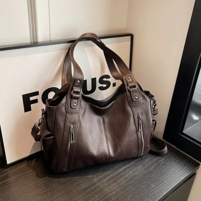 Sac à main en cuir marron foncé avec poignées robustes et poches zippées, posé sur une table noire devant un panneau "FOCUS". Accessoire mode élégant.
