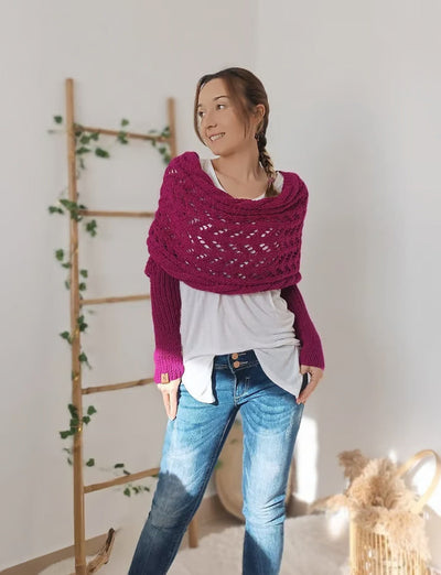 Femme portant un châle tricoté rose sur un pull blanc, posant dans un intérieur lumineux avec une échelle en bois et des plantes en arrière-plan.