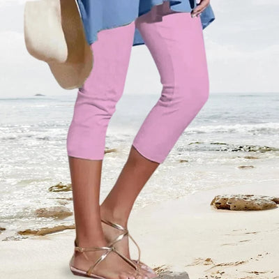 Femme en pantacourt rose et sandales dorées marchant sur une plage ensoleillée, tenant un chapeau de paille. Mode estivale, tenue décontractée bord de mer.