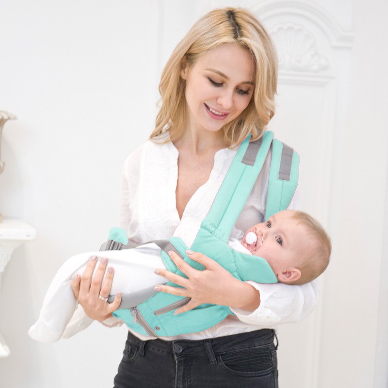 Femme souriante portant un bébé dans un porte-bébé turquoise, intérieur lumineux. Mots-clés: porte-bébé, maternité, confort, sécurité.