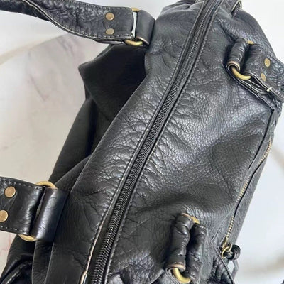 Sac à main en cuir noir texturé avec fermetures éclair dorées et poignées renforcées, posé sur une surface en marbre blanc. Accessoire mode élégant.
