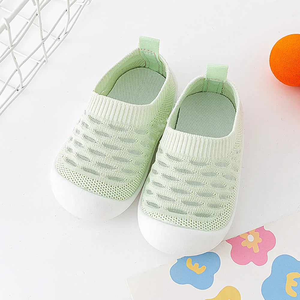 Chaussures bébé vertes en maille respirante, semelle blanche antidérapante, posées sur fond blanc avec jouet orange et papier coloré.