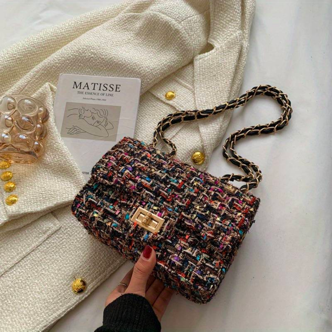 Sac à main en tweed multicolore avec chaîne dorée, posé sur un manteau blanc texturé, à côté d'un livre "Matisse" et d'un verre d'eau.