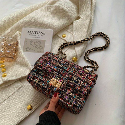 Sac à main en tweed multicolore avec chaîne dorée, posé sur un manteau blanc texturé, à côté d'un livre "Matisse" et d'un verre d'eau.
