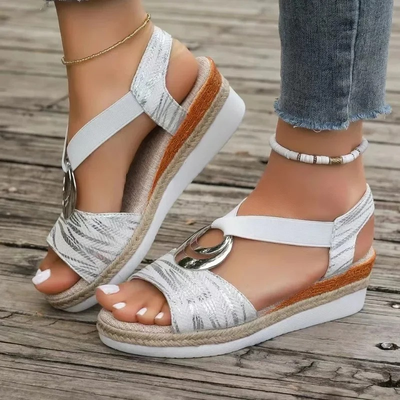 Sandales compensées blanches élégantes avec motif argenté, boucle décorative, portées avec un jean et des bracelets, sur fond de plancher en bois.