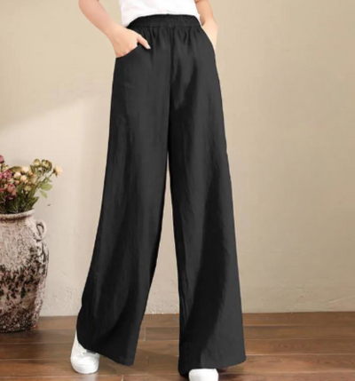Pantalon large noir femme, taille haute, style décontracté, avec poches, porté avec des baskets blanches, vase décoratif en arrière-plan.
