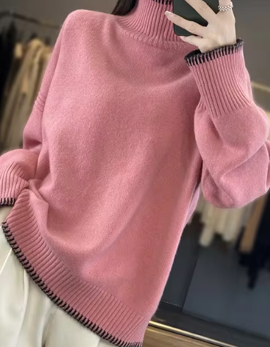 Pull rose à col roulé avec bordures contrastées noires, manches longues, porté par une personne prenant un selfie. Mode automne-hiver tendance.