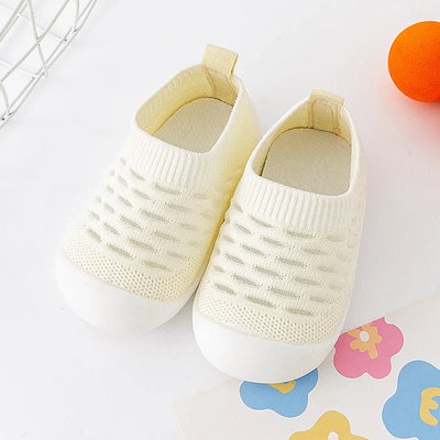 Chaussons bébé en tricot blanc, semelle antidérapante, posés sur fond blanc avec jouet orange et papier coloré. Confort et sécurité pour nourrissons.