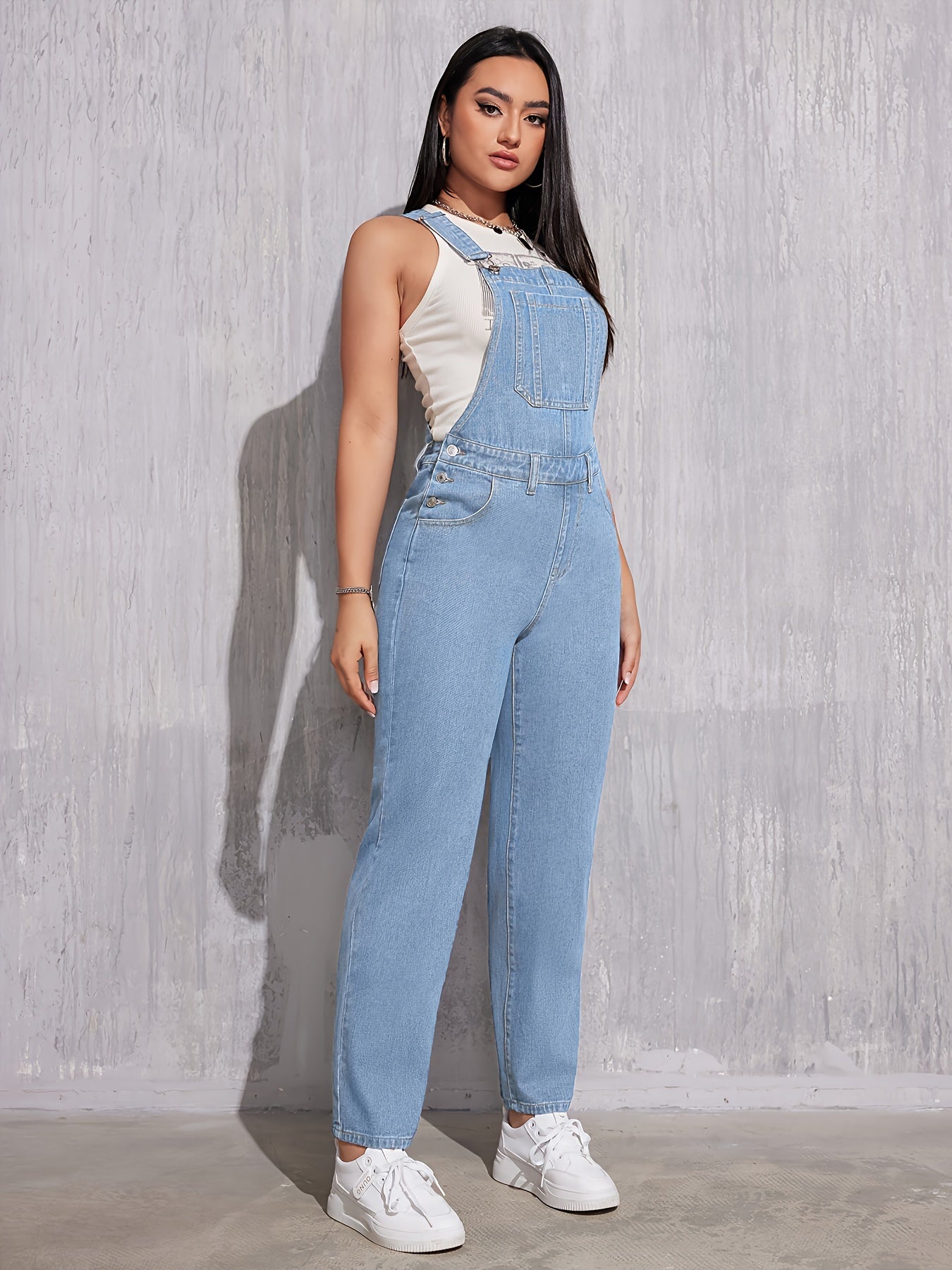 Femme portant une salopette en jean bleu clair, t-shirt blanc et baskets blanches, posant contre un mur texturé gris. Mode décontractée tendance.