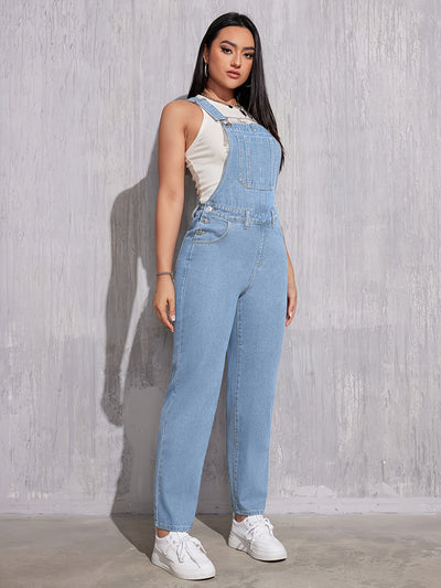 Femme portant une salopette en jean bleu clair, t-shirt blanc et baskets blanches, posant contre un mur texturé gris. Mode décontractée tendance.