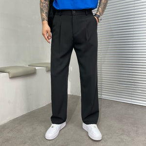 Homme en pantalon noir élégant, chemise bleue, baskets blanches, debout sur sol gris, devant mur moderne. Mode masculine, style décontracté.