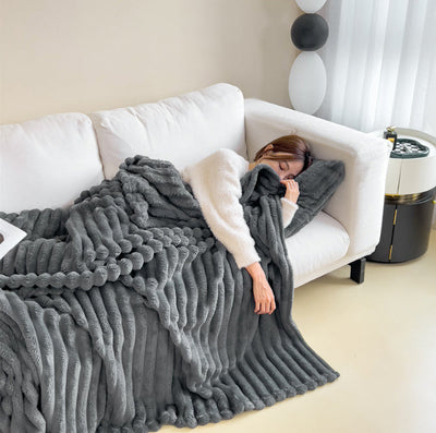 Femme endormie sur canapé blanc, enveloppée dans une couverture en fausse fourrure grise. Décor moderne, ambiance cosy et relaxante.