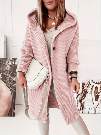 Femme portant un manteau rose en tricot avec capuche, sac blanc à bandoulière, posant devant des affiches. Mode automne-hiver, style décontracté.