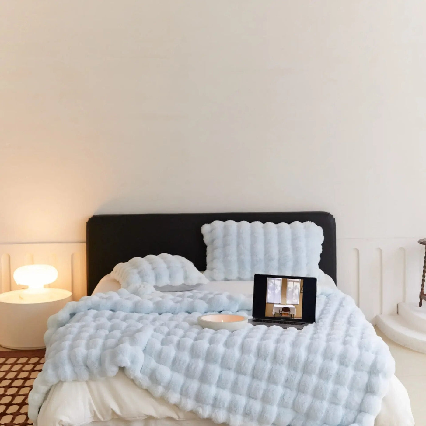Lit moderne avec couverture texturée bleue, tablette affichant une image, lampe design sur table de chevet, ambiance minimaliste et cosy.