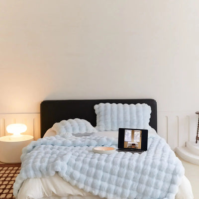 Lit moderne avec couverture texturée bleue, tablette affichant une image, lampe design sur table de chevet, ambiance minimaliste et cosy.