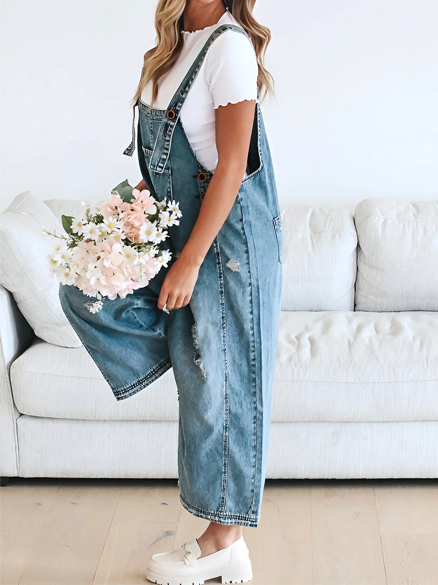 Femme en salopette en jean tenant un bouquet de fleurs roses, debout devant un canapé blanc. Mode décontractée, style printanier, intérieur moderne.