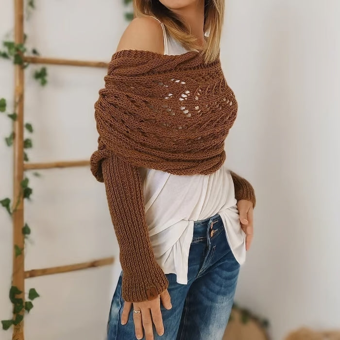 Femme portant un pull tricoté marron, style bohème, avec épaules dénudées, sur un fond de mur blanc et échelle en bois décorée de plantes.