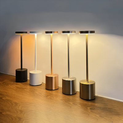 Cinq lampes de table modernes en métal de différentes couleurs (noir, blanc, cuivre, argent, or) alignées sur un sol en bois, éclairage LED.